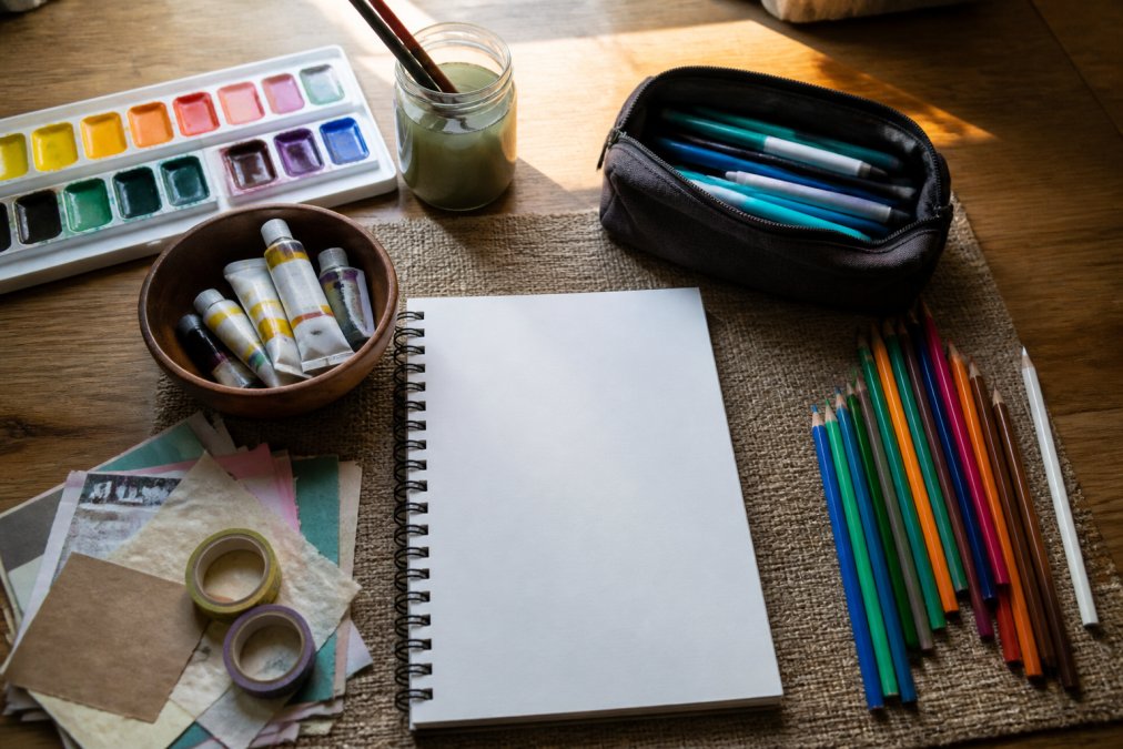 Flat lay of beginner art supplies including watercolors, gel pens, colored pencils, sketchbook, and mixed media materials arranged on a wooden table with natural window lighting