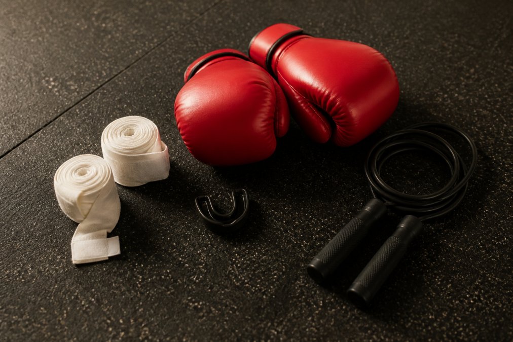 A flat lay of essential boxing gear on a gym floor: white hand wraps, red boxing gloves, a black mouthguard, and a jump rope neatly arranged with soft natural lighting