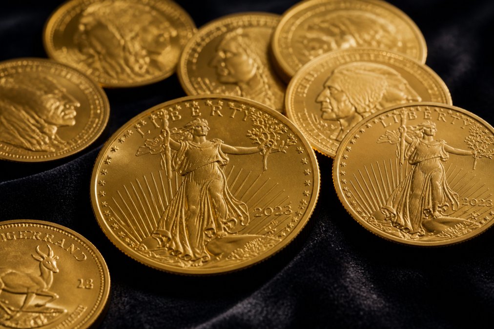 Close-up of several gold coins arranged on a dark velvet surface, including a visibly stamped American Gold Eagle coin with the date and mint mark clearly visible, soft warm studio lighting catching the luster of the metal