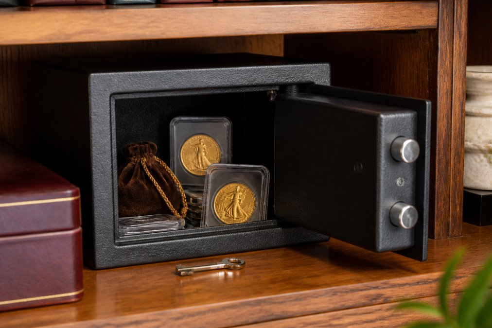 A small open home safe on a wooden shelf containing gold coins in protective clear plastic slabs and a velvet pouch, with a key resting beside it on the shelf, warm ambient lighting