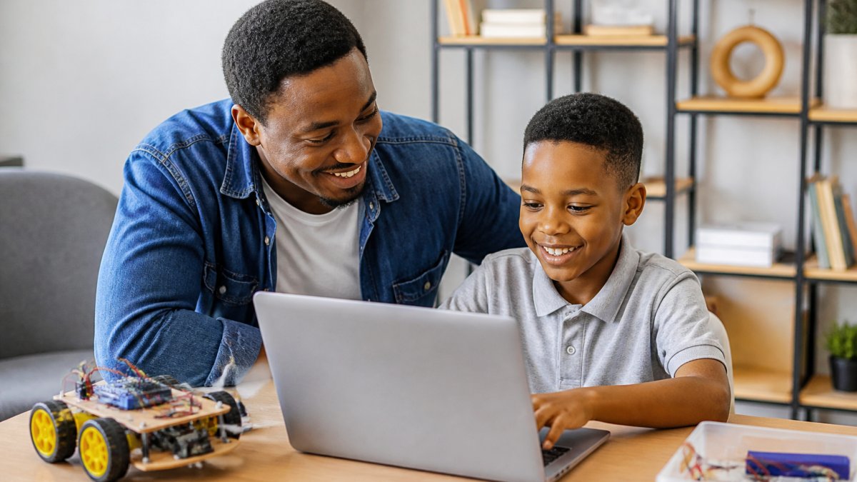 Child sitting at a desk with a laptop, engaged and smiling, with a parent nearby offering encouragement in a bright, organised home learning space