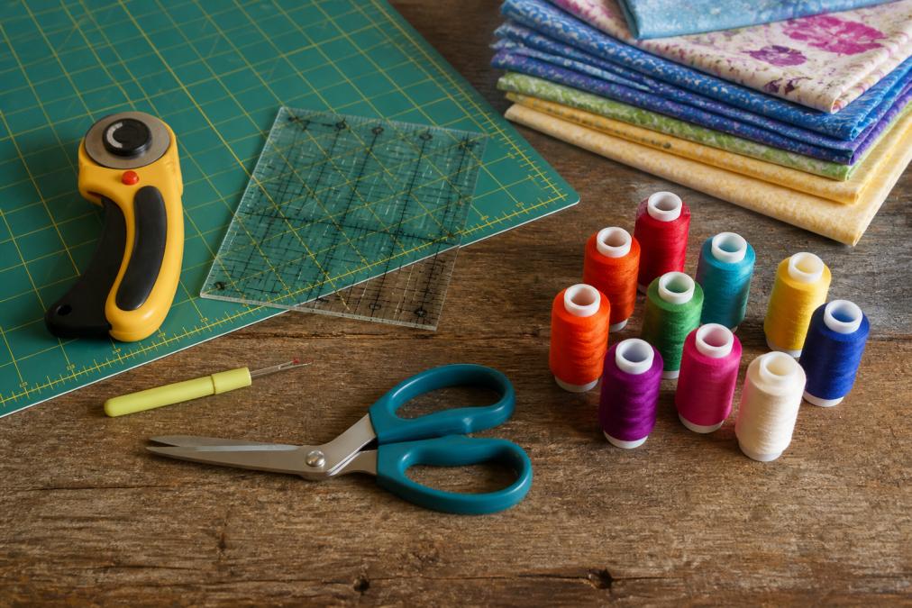 Flat lay of beginner quilting tools on a worn wooden table: a rotary cutter, self-healing mat, acrylic ruler, seam ripper, fabric scissors, and colorful spools of thread arranged neatly in warm natural light