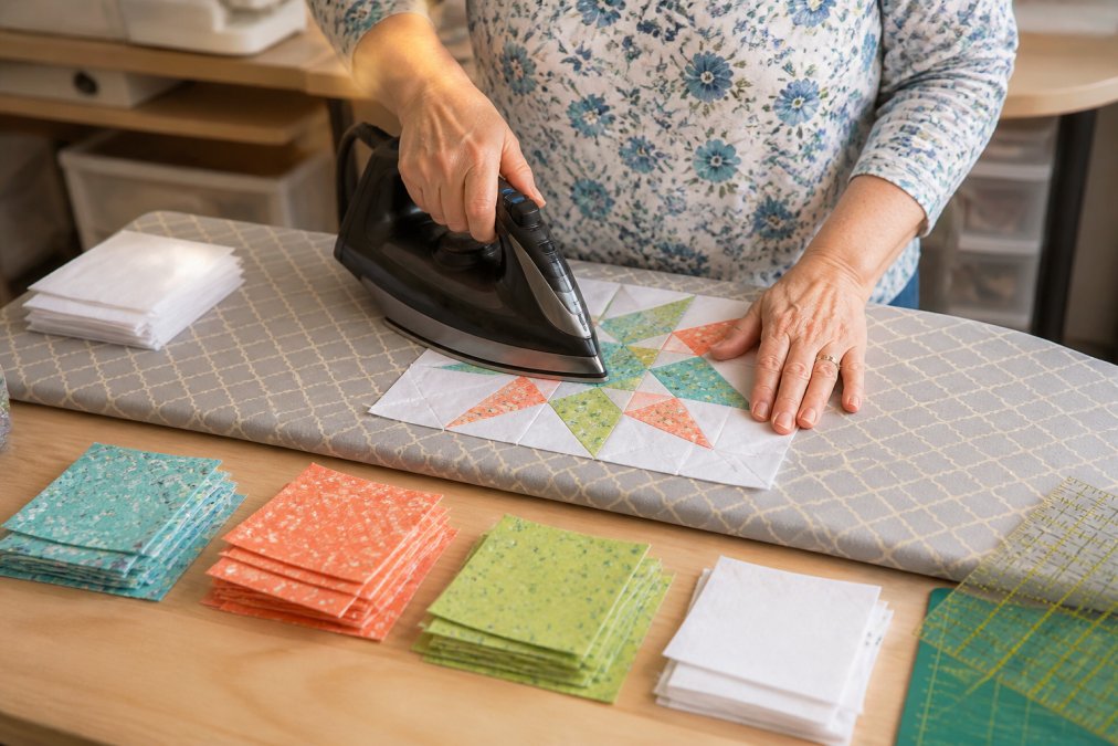 Close-up of a woman's hands pressing a freshly pieced quilt block on an ironing board, colorful quilting cotton fabric squares arranged nearby on a light wooden surface, warm indoor lighting