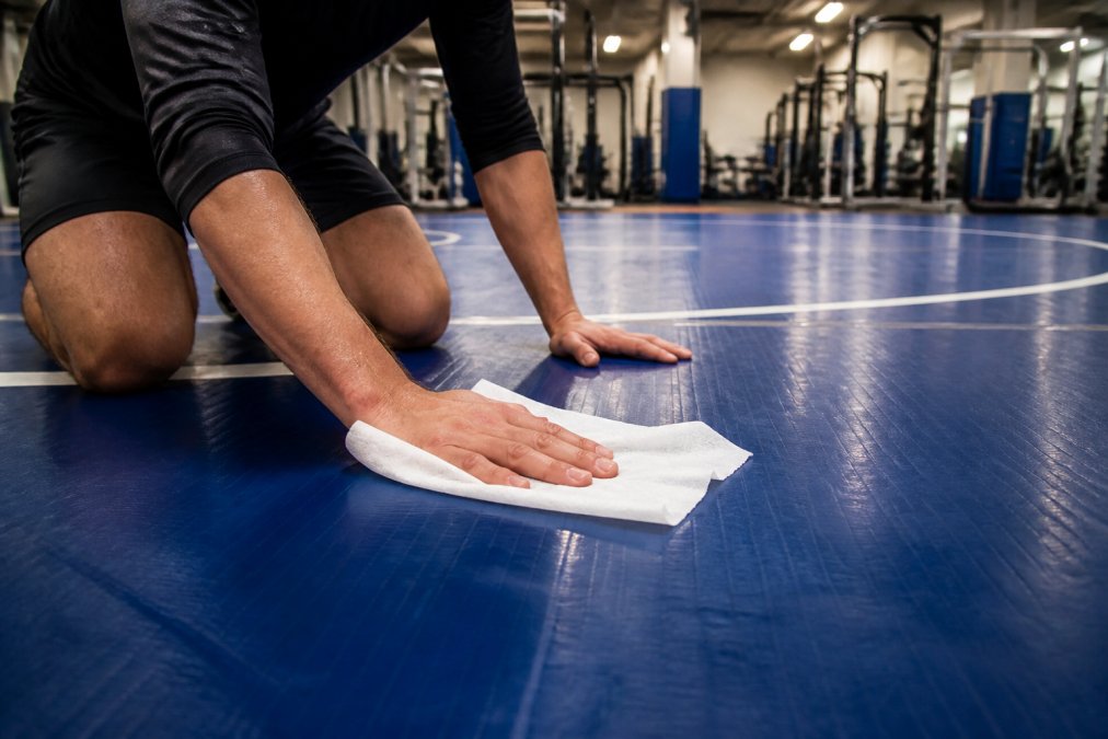Close-up of an athlete's hands wiping down a blue wrestling mat with a large antibacterial wipe, gym equipment visible and slightly blurred in the background under fluorescent lighting