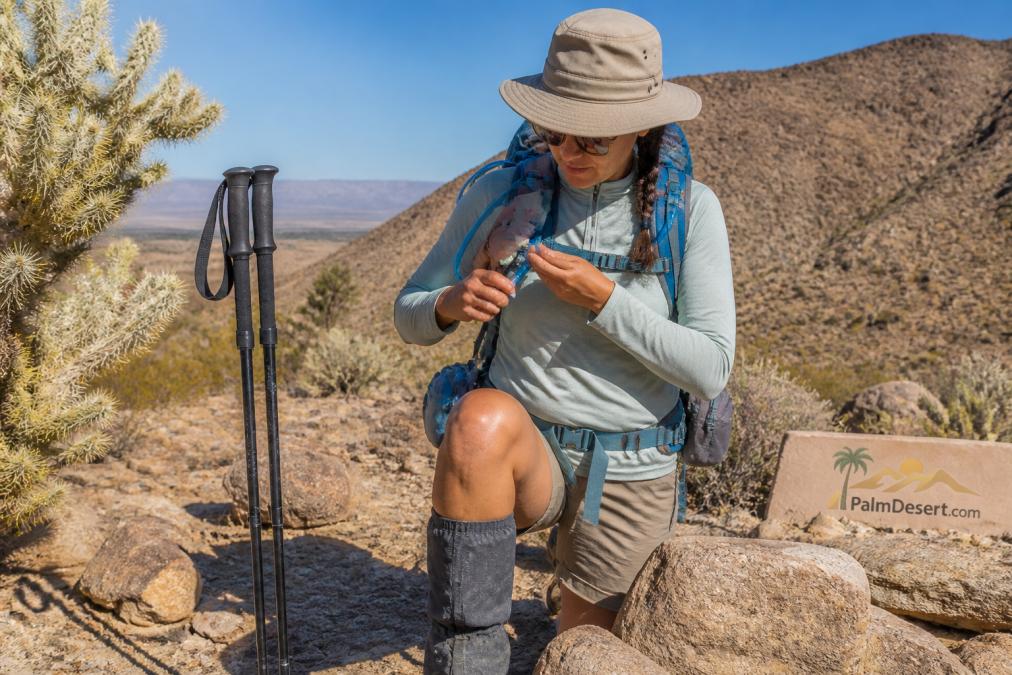 A hiker in moisture-wicking long sleeves and a broad-brimmed hat adjusting a hydration bladder on a desert trail, with trekking poles and gaiters visible, bright sunlight and rocky terrain in the background