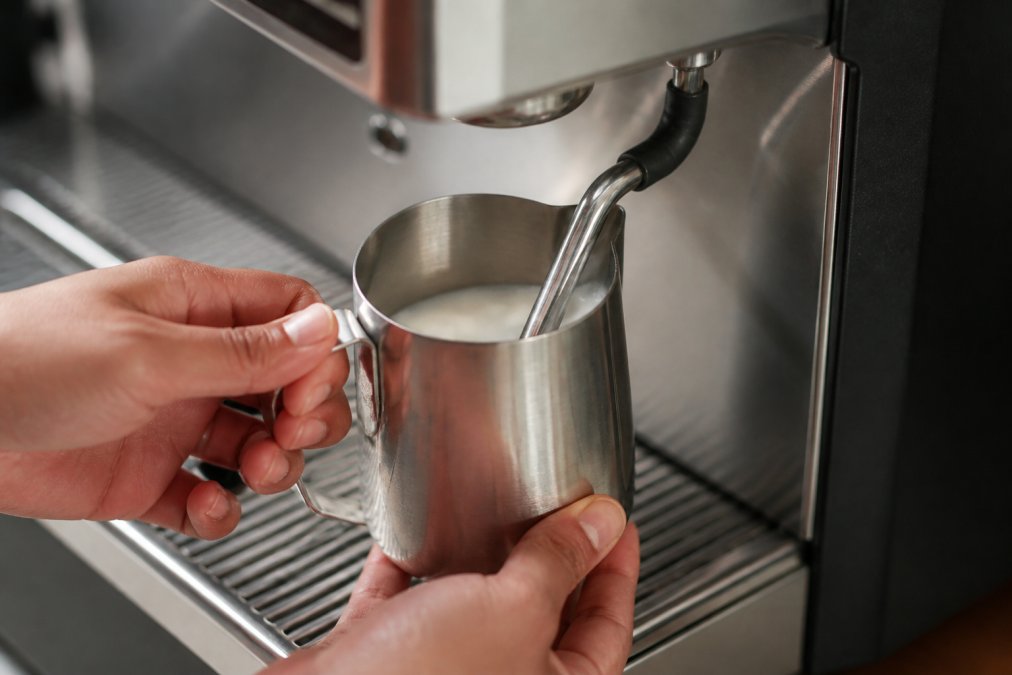Close-up of hands positioning a stainless steel frothing pitcher under a steam wand on an espresso machine, showing proper 45-degree angle with steam nozzle just below milk surface