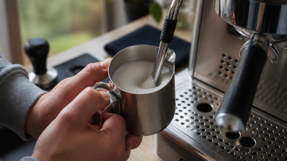 Close-up of hands holding a stainless steel milk pitcher under a steam wand on a home espresso machine, wand tip positioned just below the milk surface with a visible whirlpool forming