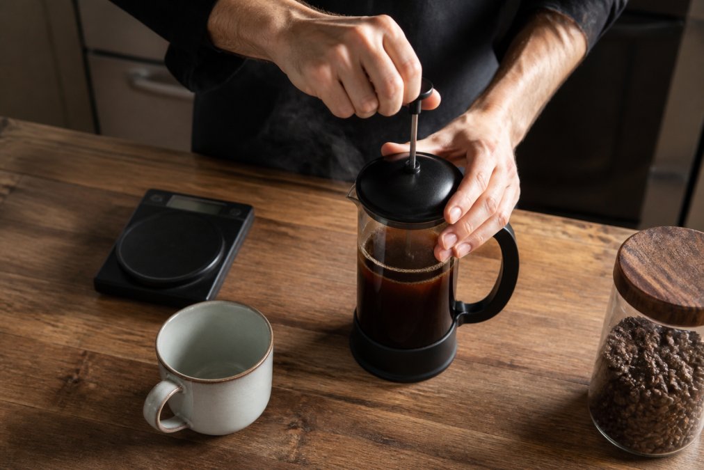 Overhead shot of hands pressing the plunger down on a French press filled with dark coffee, steam rising, on a wooden kitchen counter with a ceramic mug and digital scale nearby