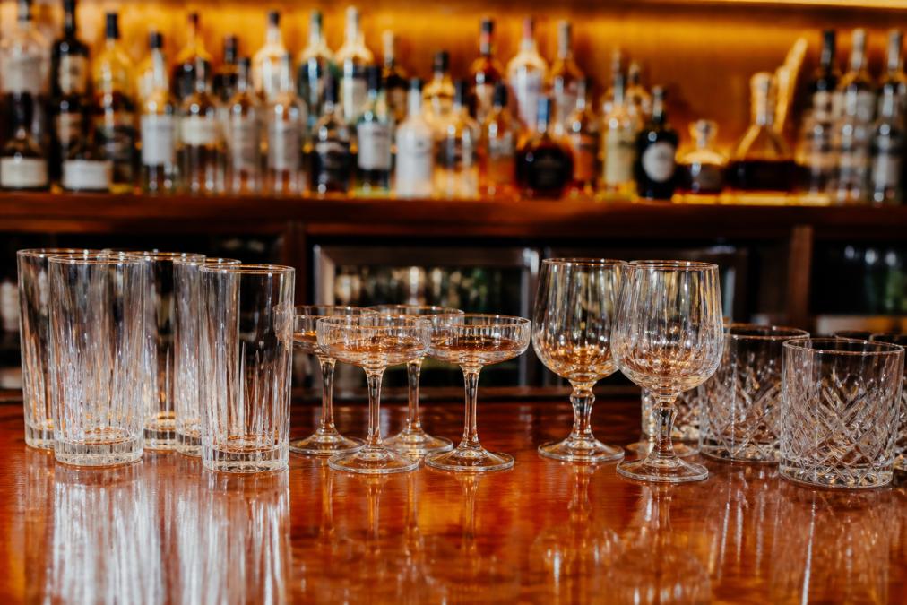 Selection of bar glassware including highball glasses, coupe glasses, and specialty cocktail glasses arranged on a polished wooden bar counter with soft warm lighting, showing reflections of the bottles behind