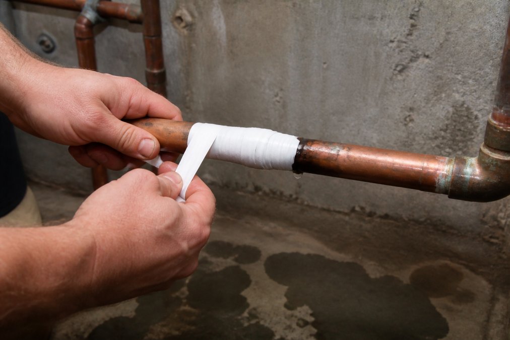 Hands wrapping waterproof plumbing repair tape around a burst pipe section in a basement, demonstrating the overlapping wrapping technique with clear visibility of the damaged area