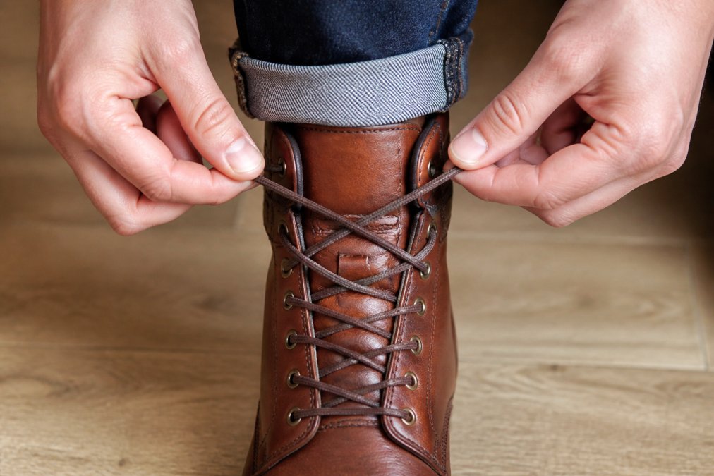 Close-up of hands demonstrating heel-lock lacing technique on a brown leather boot, showing the crisscross pattern at the ankle and heel area with the lace creating a secure loop