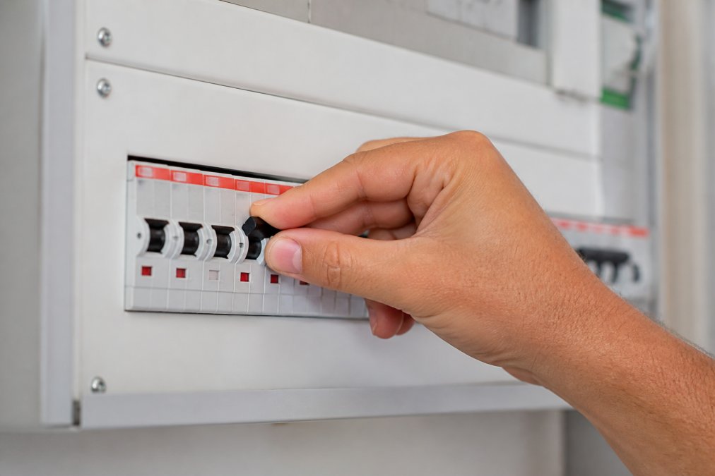 A person's hand firmly flipping a single circuit breaker switch to the 'on' position inside a modern residential switchboard, with other breakers clearly labelled