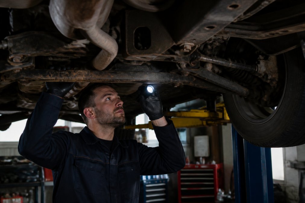 Mechanic using a flashlight to inspect vehicle frame rails to stop car frame rust during surface preparation.