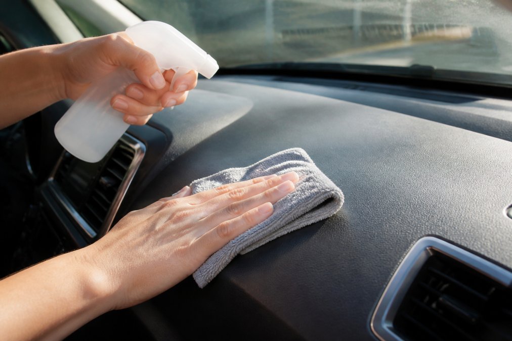 Close-up of hands cleaning a car dashboard with a microfiber cloth and cleaning solution, showing removal of dust and residue from the mounting surface in natural sunlight