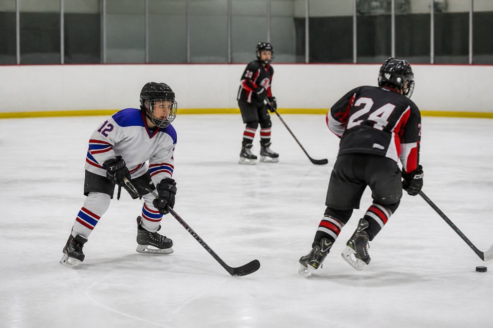 A youth hockey player on ice with head up and eyes scanning toward the puck carrier, holding a defensive stance while other players move around them