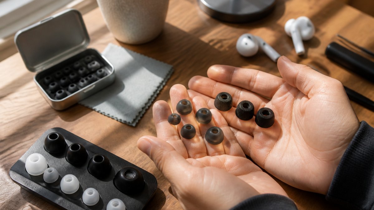 Close-up of a person's hands holding three different earbud tip sizes (small, medium, large silicone and foam tips) over a wooden desk in soft daylight