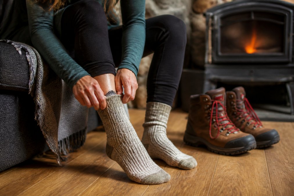 Close-up of a person's feet wearing thick alpaca wool socks indoors, with warm winter boots positioned nearby on a wooden floor, natural indoor lighting