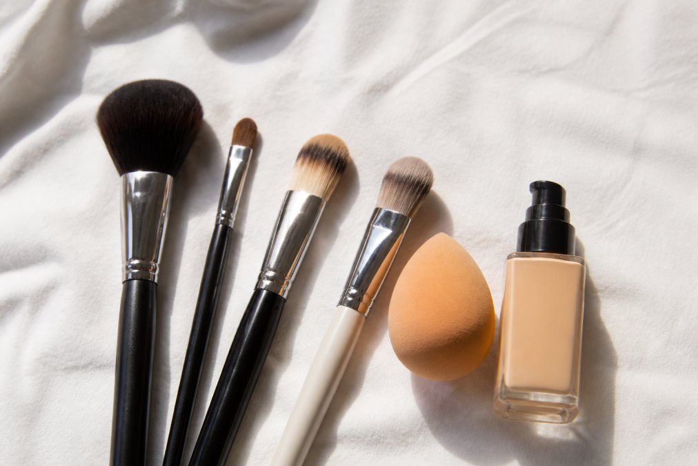 Flat lay of professional makeup brushes, a damp beauty sponge, and a foundation bottle arranged on a soft white linen surface in bright, even daylight