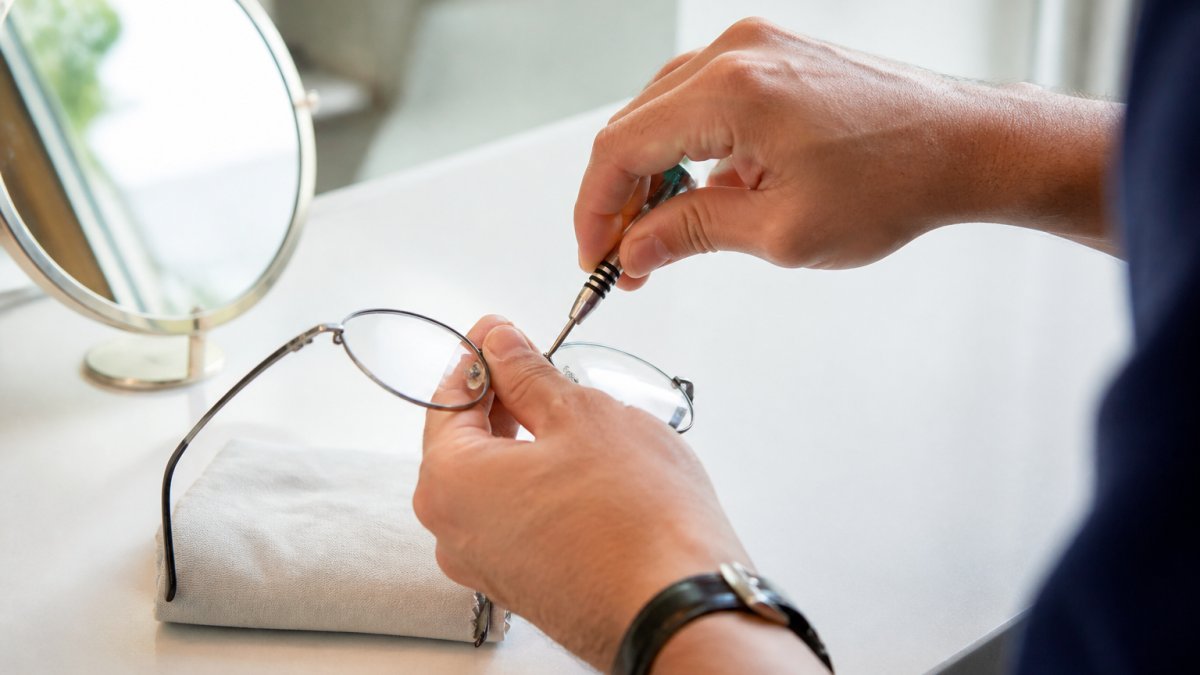 Close-up of a person's hands using a small screwdriver to gently adjust the silicone nose pads on a pair of metal eyeglasses, with a mirror and soft cloth on a clean table in a bright room