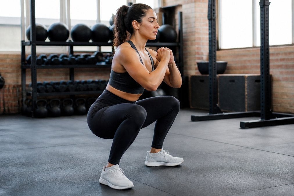 Woman performing a deep squat in a gym wearing high-waisted leggings, demonstrating proper fit and no slippage at the waistband