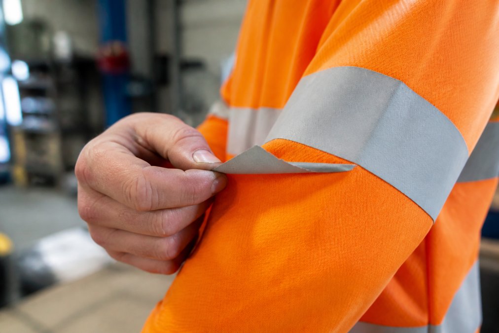 Close-up of a worker's hands inspecting the edges of reflective tape on an orange hi vis shirt sleeve, checking for lifting or peeling under bright workshop lighting