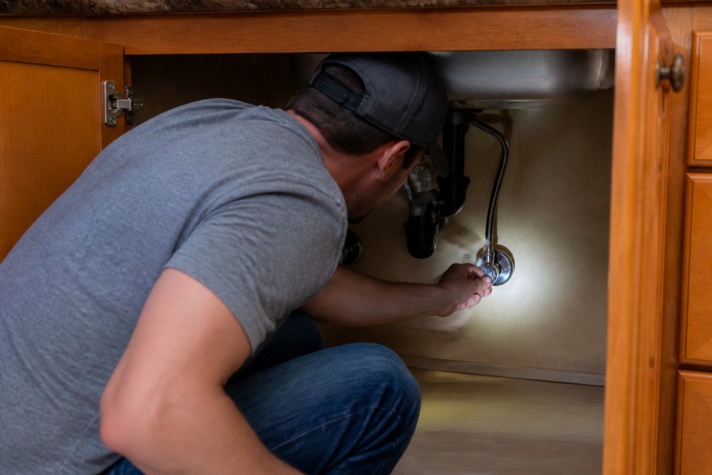 Person crouching under a kitchen sink, turning a chrome shut-off valve by hand with cabinet doors open and pipes visible in the background, illuminated by a flashlight