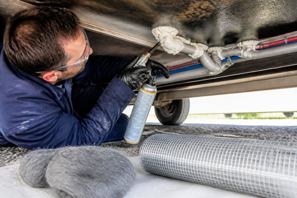 Pest control professional in protective gloves and safety glasses carefully applying spray foam to gaps around plumbing penetrations in a trailer underbelly, with steel wool and hardware cloth materials visible on the work surface