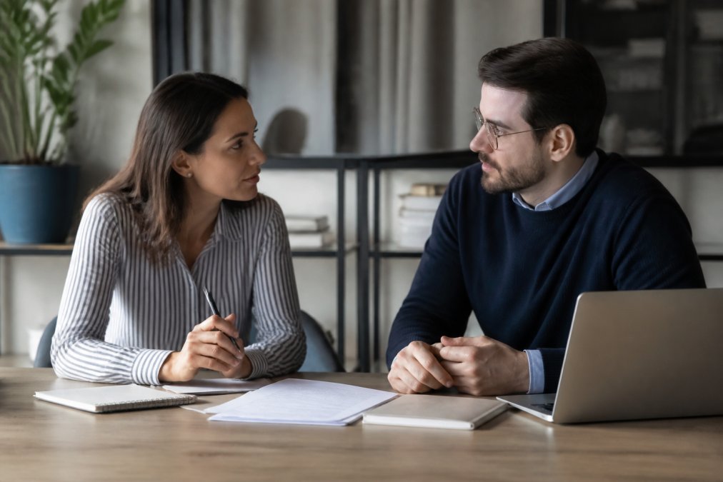Manager and team member having a focused one-on-one conversation at a desk, both engaged and making direct eye contact, with notes and a laptop visible on the surface between them
