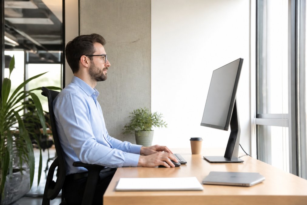 Professional working at desk with monitor positioned at eye level, perpendicular to natural window light, showing proper ergonomic posture and monitor tilt angle