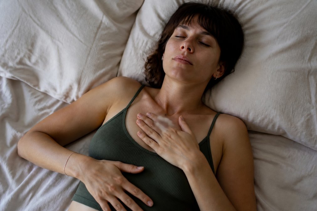 A calm, softly lit bedroom scene where a person lies on a bed with eyes closed, hands resting gently on their chest and abdomen, embodying a grounding body-awareness practice