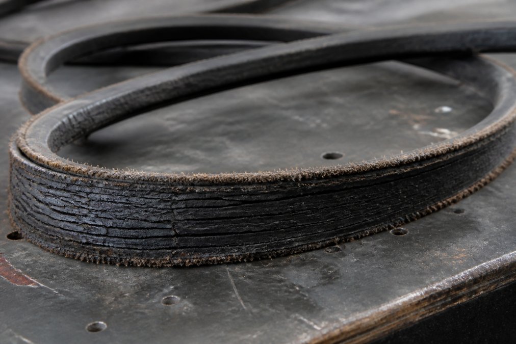 Close-up of a worn industrial V-belt laid on a workbench under bright workshop lighting, showing visible transverse cracking, glazed contact surfaces, and fraying along the belt edges