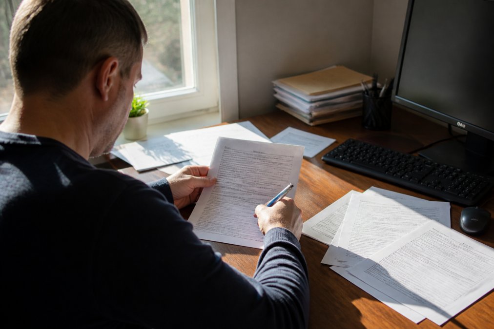 Person sitting at a wooden desk with legal documents spread out, a pen in hand, computer monitor showing a form, natural window lighting illuminating the paperwork