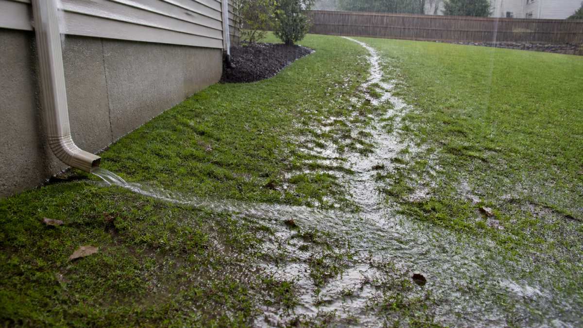 Backyard sloping gently away from house foundation during light rain, showing water runoff flowing downhill across grass toward yard perimeter