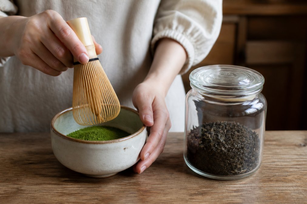 Hands holding a traditional matcha whisk suspended over a ceramic bowl containing vibrant green matcha powder, positioned next to a glass jar of loose leaf tea, showing the visual and textural contrast between the two forms