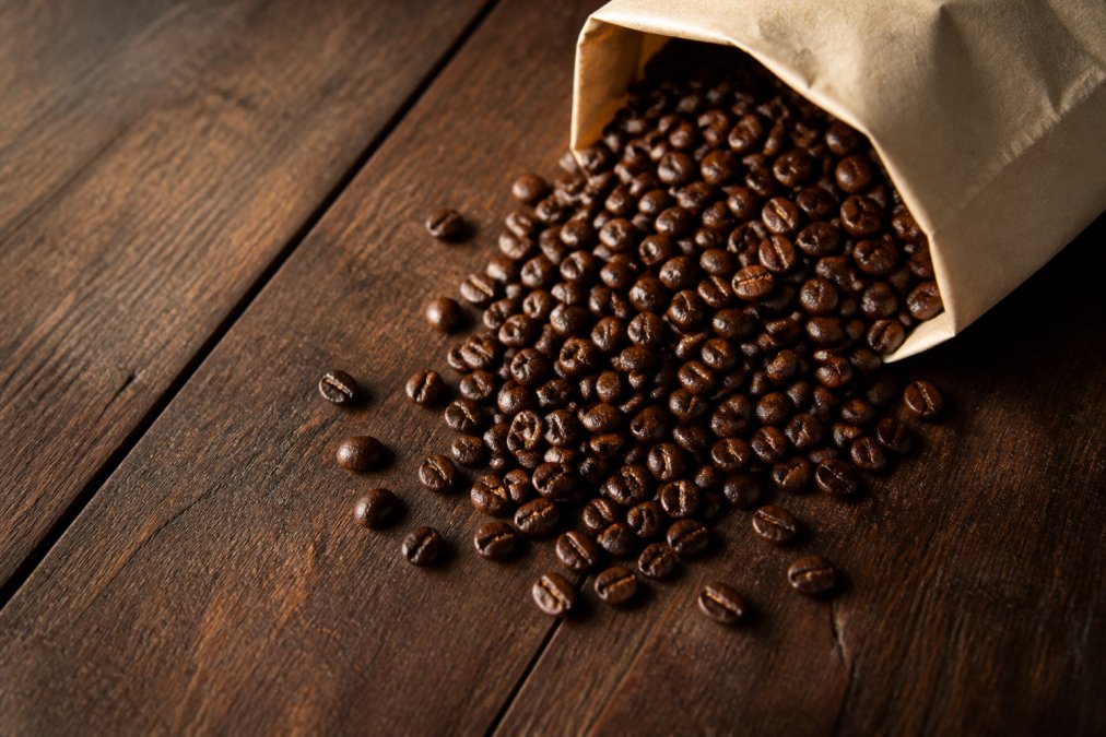 Overhead shot of freshly roasted whole coffee beans spilling from a brown paper bag onto a dark wooden table, with visible oils glistening on the beans under warm window light