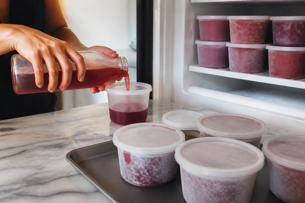 Hands carefully pouring shrub soda into freezer-safe containers, with labeled containers already frozen in background on freezer shelf
