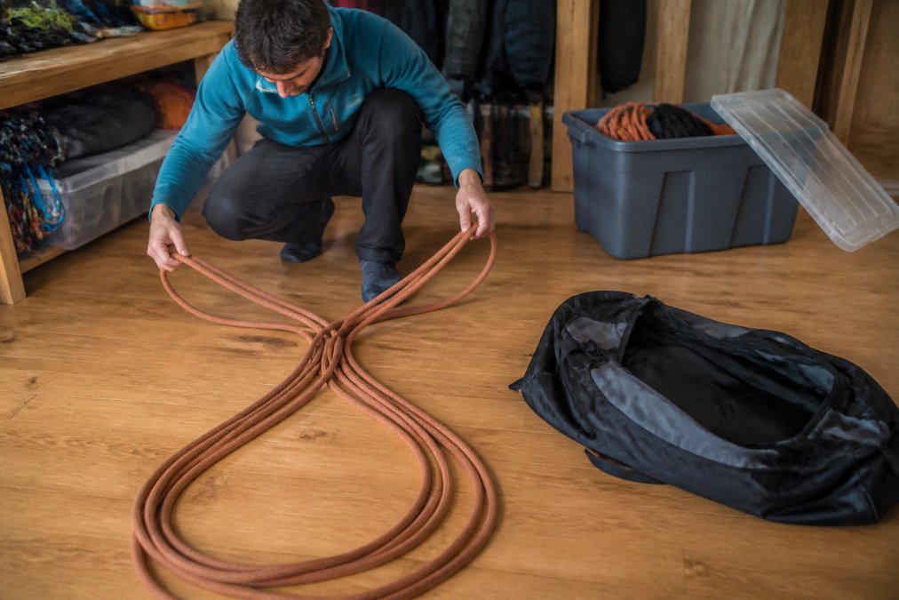 A climber coiling a dynamic climbing rope on a clean wooden floor using the butterfly method, with a rope bag and a labeled storage bin nearby in a cool, dry room