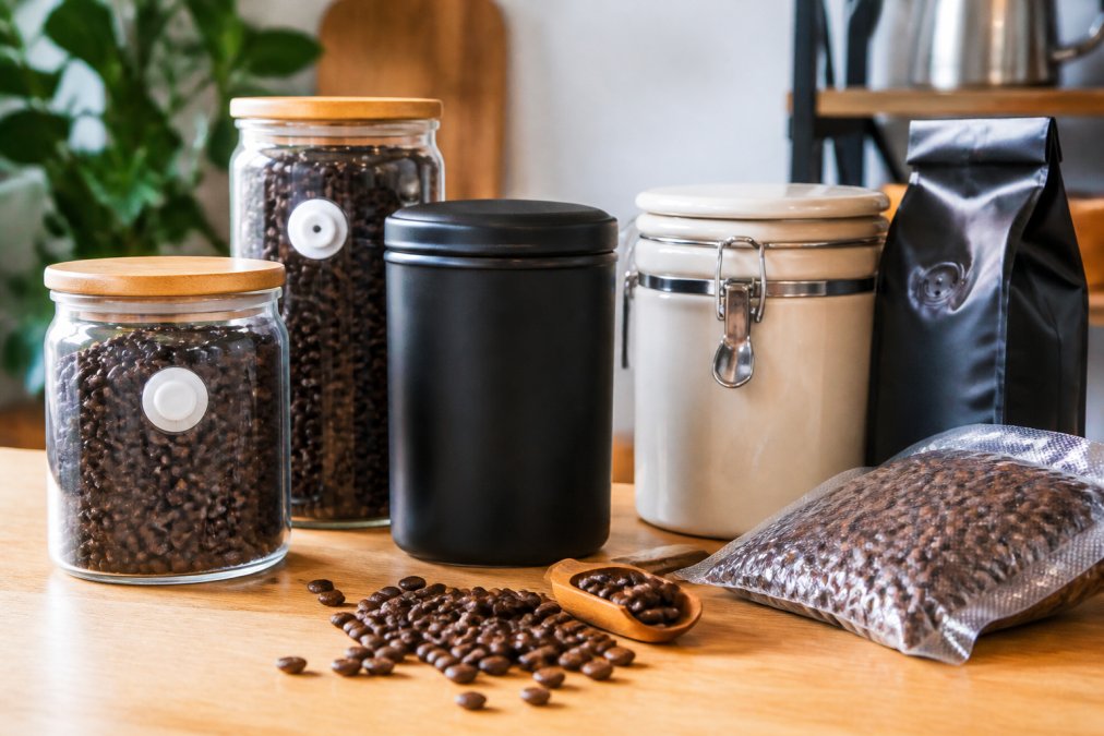 Close-up of various coffee storage containers including clear glass jars with one-way valves, opaque ceramic canisters, and vacuum-sealed bags with coffee beans scattered on a light wooden counter surface
