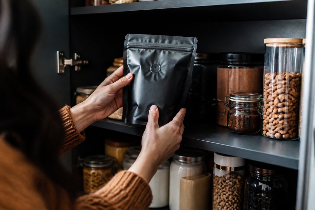 A person's hands placing a sealed coffee bag into a dark, organized pantry cabinet, with other pantry items softly blurred in the background