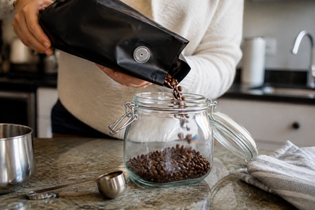 Hands pouring roasted coffee beans into an airtight glass container to store coffee beans properly.