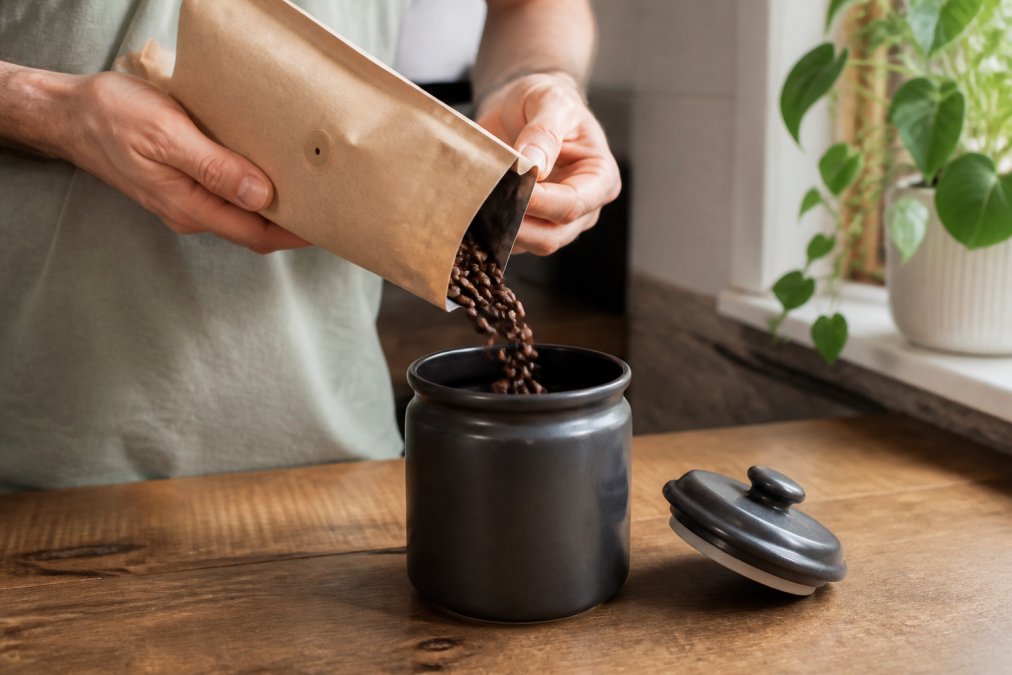 A person's hands pouring whole coffee beans from a small paper bag with a one-way valve into a dark, opaque ceramic canister with a tight-fitting lid on a kitchen counter. Soft natural light from a nearby window illuminates the beans mid-pour.