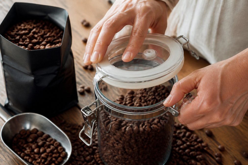 Close-up of hands sealing a premium coffee bag with an airtight container, showing the rubber gasket and one-way valve clearly visible, with whole Colombian beans scattered on a wooden surface in soft natural light