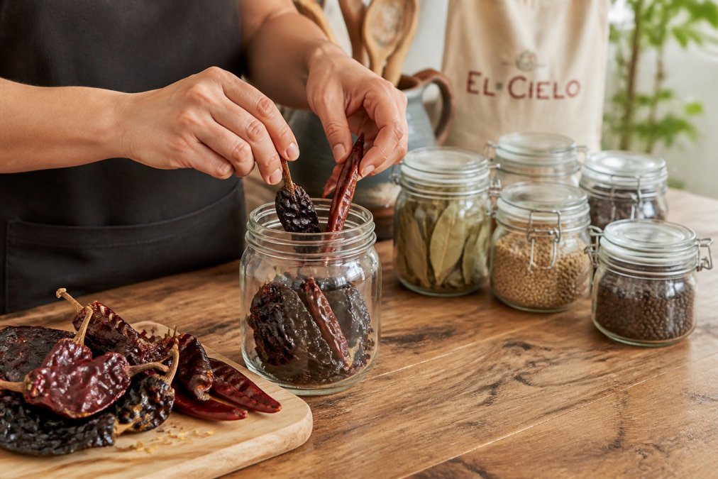 Home cook placing whole dried ancho and guajillo chillies into a glass jar to store dried chillies properly.