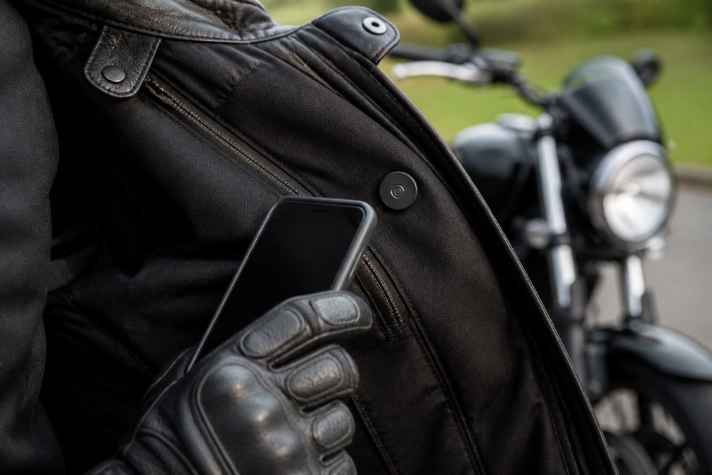 Close-up photograph of a motorcyclist's gloved hand holding a smartphone against a small NFC emergency tag attached to the inside flap of a black leather motorcycle jacket, with the bike visible out of focus behind