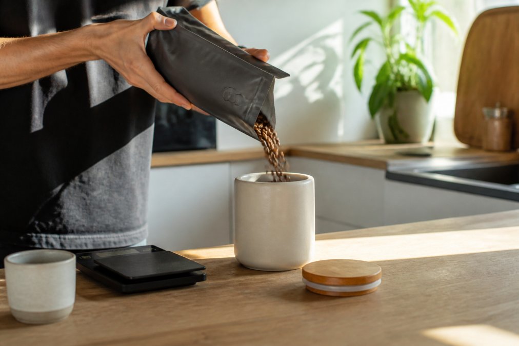 A person's hands pouring whole coffee beans from a one-way valve bag into an opaque ceramic storage canister on a kitchen counter, with a digital scale and a mug nearby in warm morning light