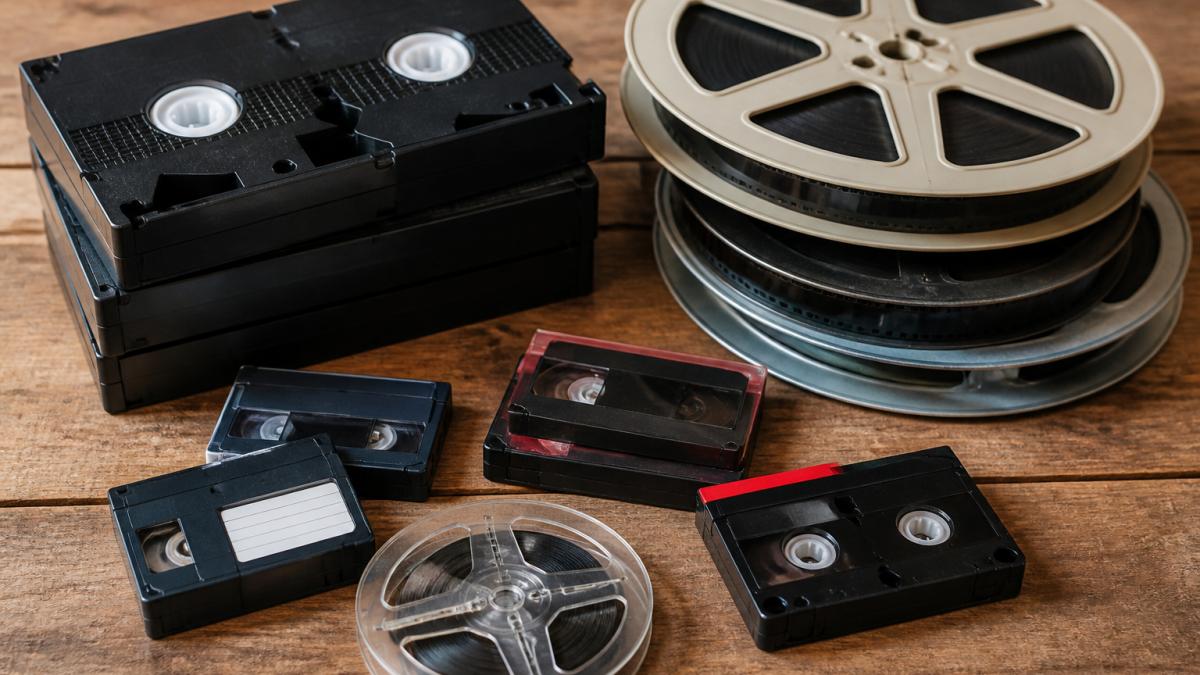 Collection of vintage media formats arranged on wooden surface including VHS tapes, film reels, 8mm cassettes, and old video cassettes showing variety of formats families preserve