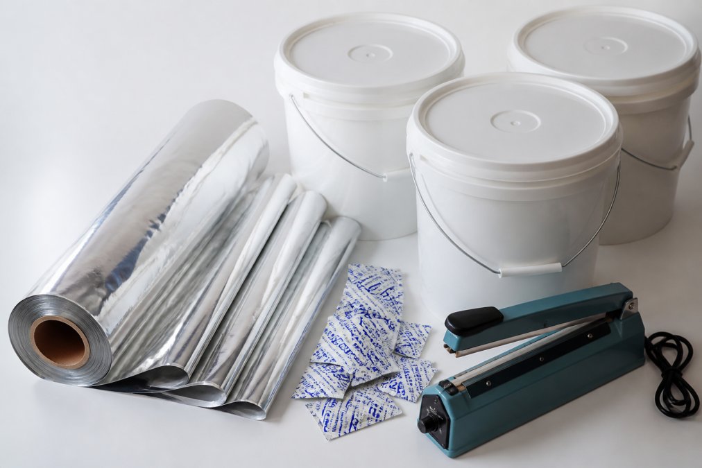 Flat lay of rice storage supplies arranged on white background: rolled mylar bags, food-grade plastic buckets with lids, small packets of oxygen absorbers, and a handheld heat sealing tool