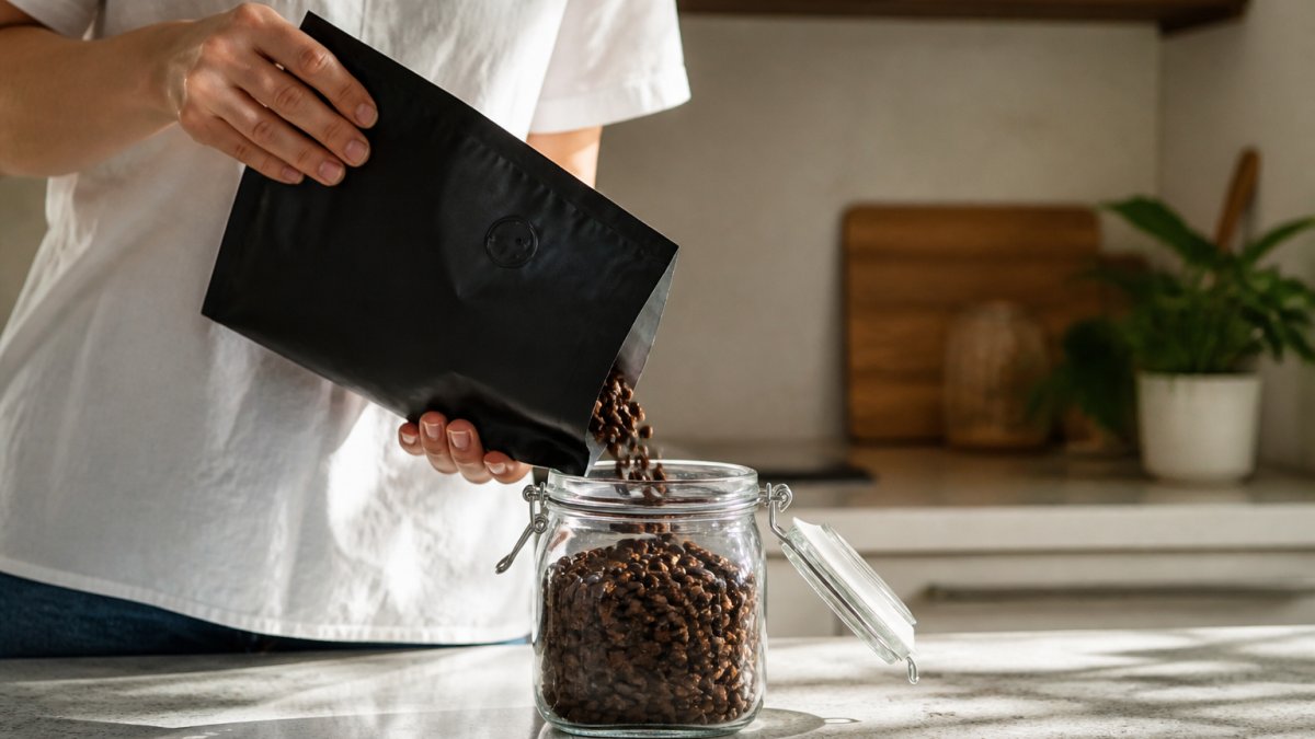 A person placing a bag of roasted coffee beans into an airtight glass jar on a kitchen counter, with a one-way valve visible on the bag, soft morning light