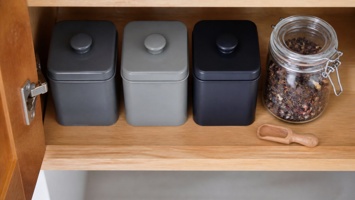Overhead shot of an open kitchen cabinet shelf holding three opaque airtight tea tins next to a glass jar of loose leaf tea, soft natural light, a small wooden scoop resting beside them