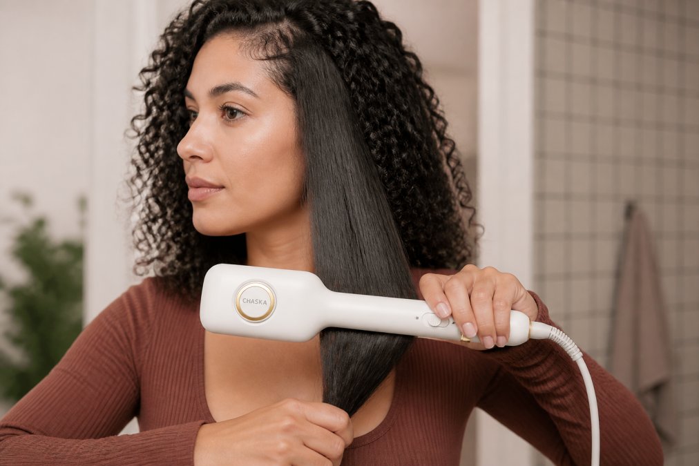 Woman straightening a section of thick curly hair with a flat iron, demonstrating smooth downward pass-through motion with consistent tension, showing the hair transitioning from curly to straight through the plates